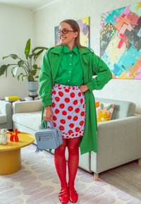 Grønn oversized jakke over en lys grønn skjorte, mønstrete skjørt med røde sirkler, røde strømpebukser, røde sko og en lyseblå veske.