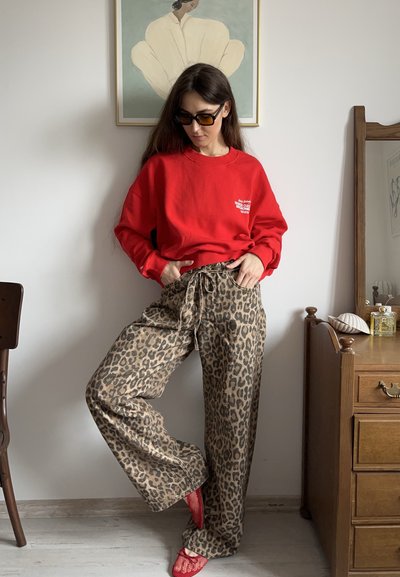 Sudadera roja con estampado gráfico, combinada con pantalones de corte suelto de estampado de leopardo. Completado con zapatillas rojas de malla sin cordones. Muebles de madera al fondo.