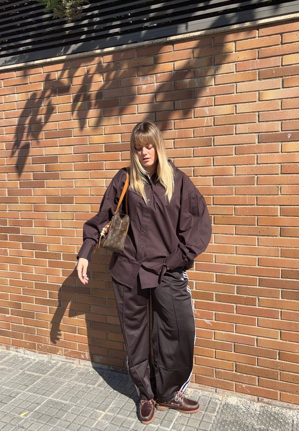 Una mujer vestida de oscuro está de pie al sol junto a una pared de ladrillo y lleva un pequeño bolso estampado al hombro.
