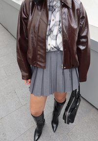 Giacca di pelle lucida marrone sopra una camicia a quadri con bottoni, abbinata a una gonna grigia plissettata e stivali neri con punta, al ginocchio. Borsa nera.