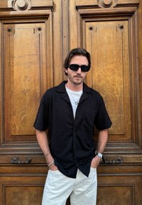 Μαύρο κοντομάνικο πουκάμισο με υφή, ενιαία τσέπη στο στήθος και άνετη εφαρμογή; συνδυασμένο με κρέμ παντελόνι και γυαλιά ηλίου.