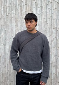 Νέος άνδρας με γκρι πουλόβερ και μαύρη τσάντα ώμου στέκεται μπροστά σε λευκό τοίχο από τούβλα με υφή, κοιτάζοντας ελαφρώς προς τα δεξιά.