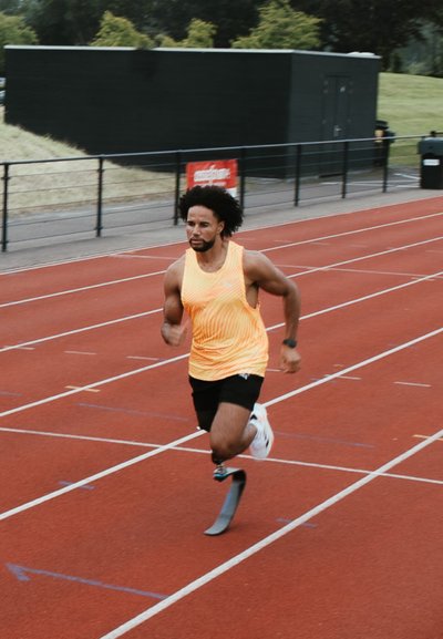 Atleta corriendo en una pista roja, vistiendo una camiseta sin mangas de color naranja brillante y pantalones cortos negros, con una pierna prostética y zapatillas de correr blancas.