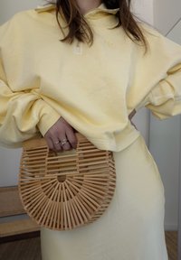 Κίτρινη ζακέτα με μισό φερμουάρ και δ�ιακριτό λογότυπο, συνδυασμένη με μια κυκλική τσάντα από μπαμπού με πλαισιωτό σχέδιο.