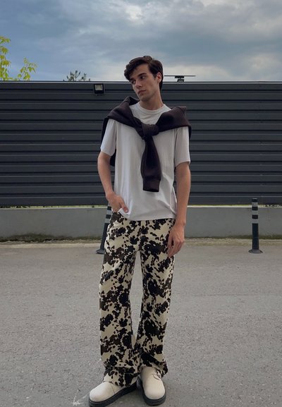 Camiseta blanca de cuello redondo, suéter marrón oscuro colgado, pantalones beige con patrón abstracto negro y zapatos color crema con suelas negras.