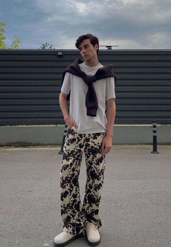 White crew-neck t-shirt, dark brown sweater draped over it, beige trousers with a black abstract pattern, and cream-coloured shoes with black soles.