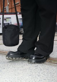Zapatos de plataforma negros con suelas de goma gruesa y texturizada, combinados con pantalones sueltos negros y un pequeño bolso cruzado negro.