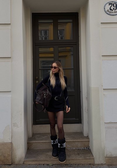 Mujer con atuendo negro, gafas de sol y botas de invierno, de pie en unas escaleras sosteniendo una gran bolsa frente a una puerta oscura de madera con paneles de vidrio.