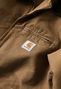 Hnědá bavlněná bunda s uzávěrem na zip, levým náprsním kapsou a nášivkou Carhartt v bílé a oranžové barvě. Viditelný texturovaný detail látky.