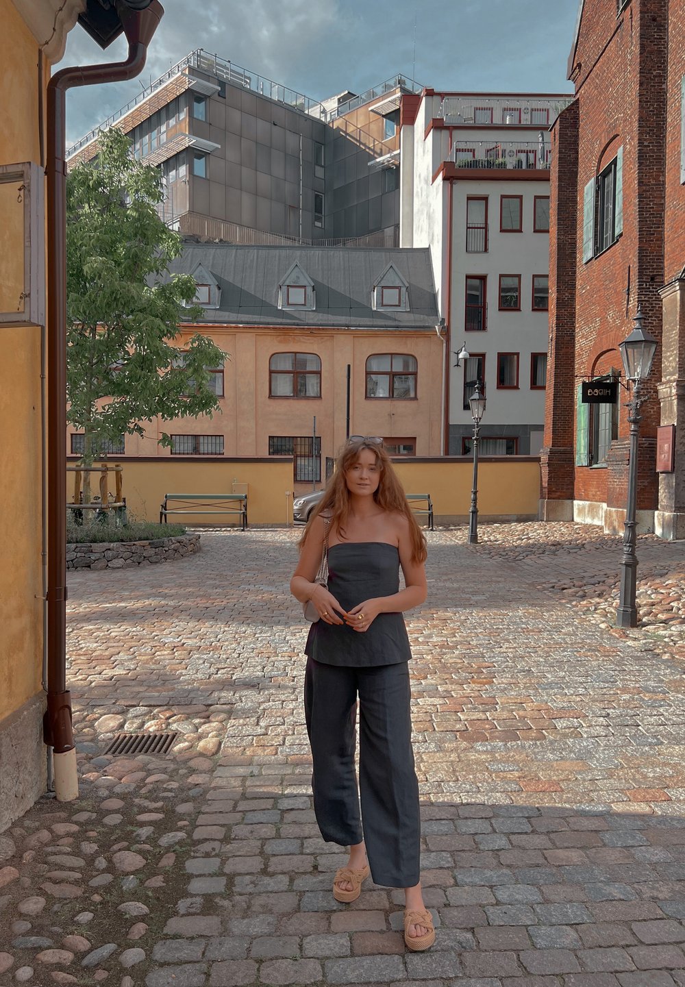 Μαύρη, χωρίς τιράντες μπλούζα και παντελόνι σε φαρδιά γραμμή, συνδυασμένα με καφέ, πλεγμένες σαγιονάρες. Πλάκα από πέτρες με κτίρια στο φόντο.