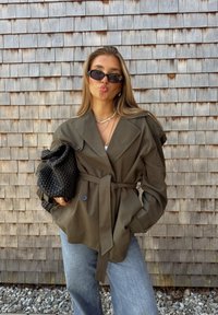Giacca belted verde oliva con ampi risvolti, dettagli con bottoni e tessuto texturizzato. Abbinata a una borsa nera intrecciata e jeans in denim chiaro.