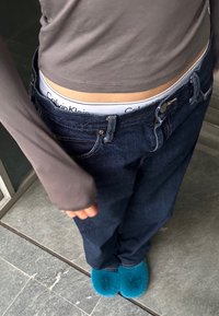 Jeans de mezclilla azul oscuro con cierre de botón y cremallera, combinados con una blusa gris de manga larga y pantuflas de felpa turquesa. Cintura de Calvin Klein visible.