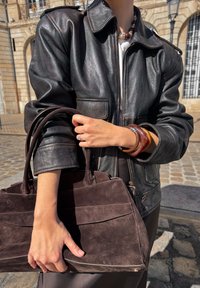 Person in black leather jacket holding dark brown suede tote bag, wearing chunky bracelets and a beaded necklace, standing on cobblestone street.