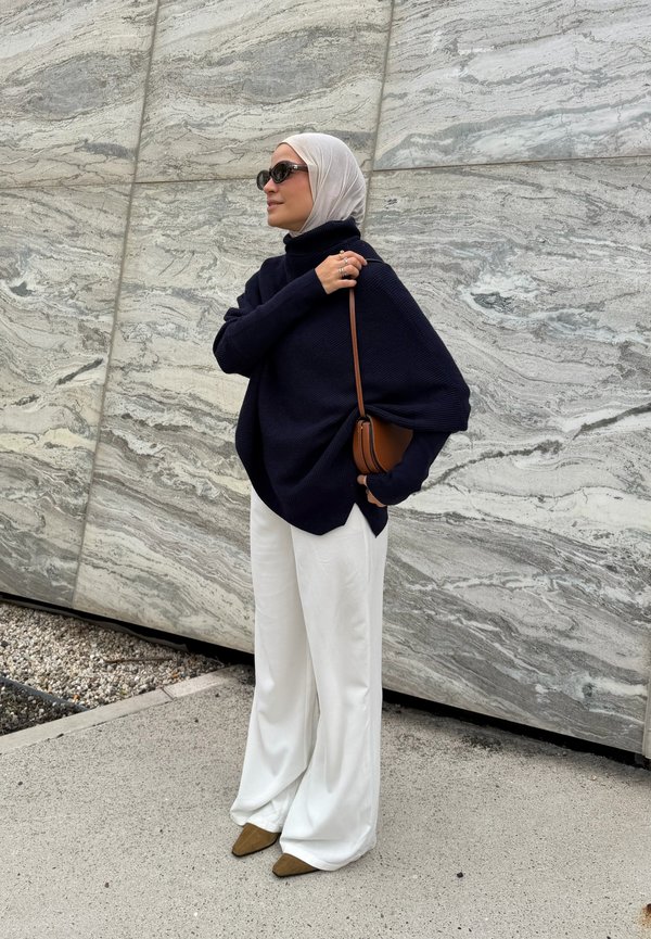 Woman wearing white hijab, black oversized sweater, white wide pants, brown shoes, and sunglasses standing against a gray marble wall.