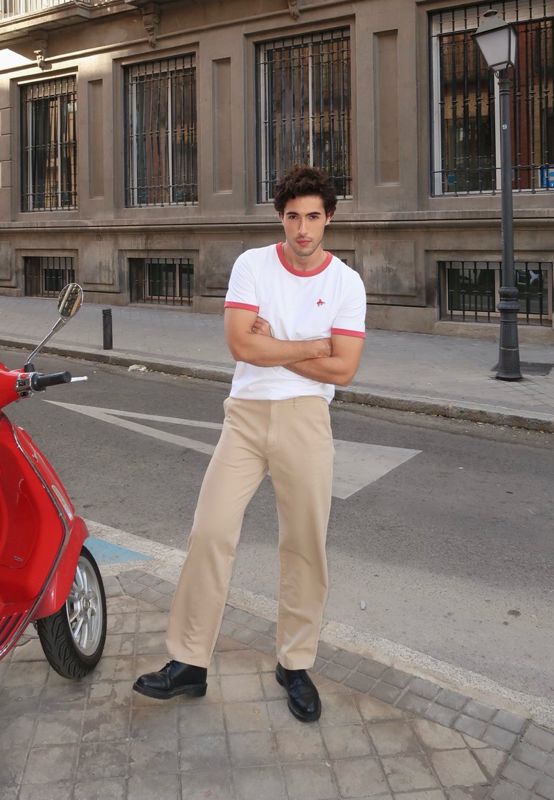 Conjunto casual com uma t-shirt branca com detalhes em vermelho, calças bege e sapatos pretos. Uma scooter vermelha está posicionada nas proximidades.