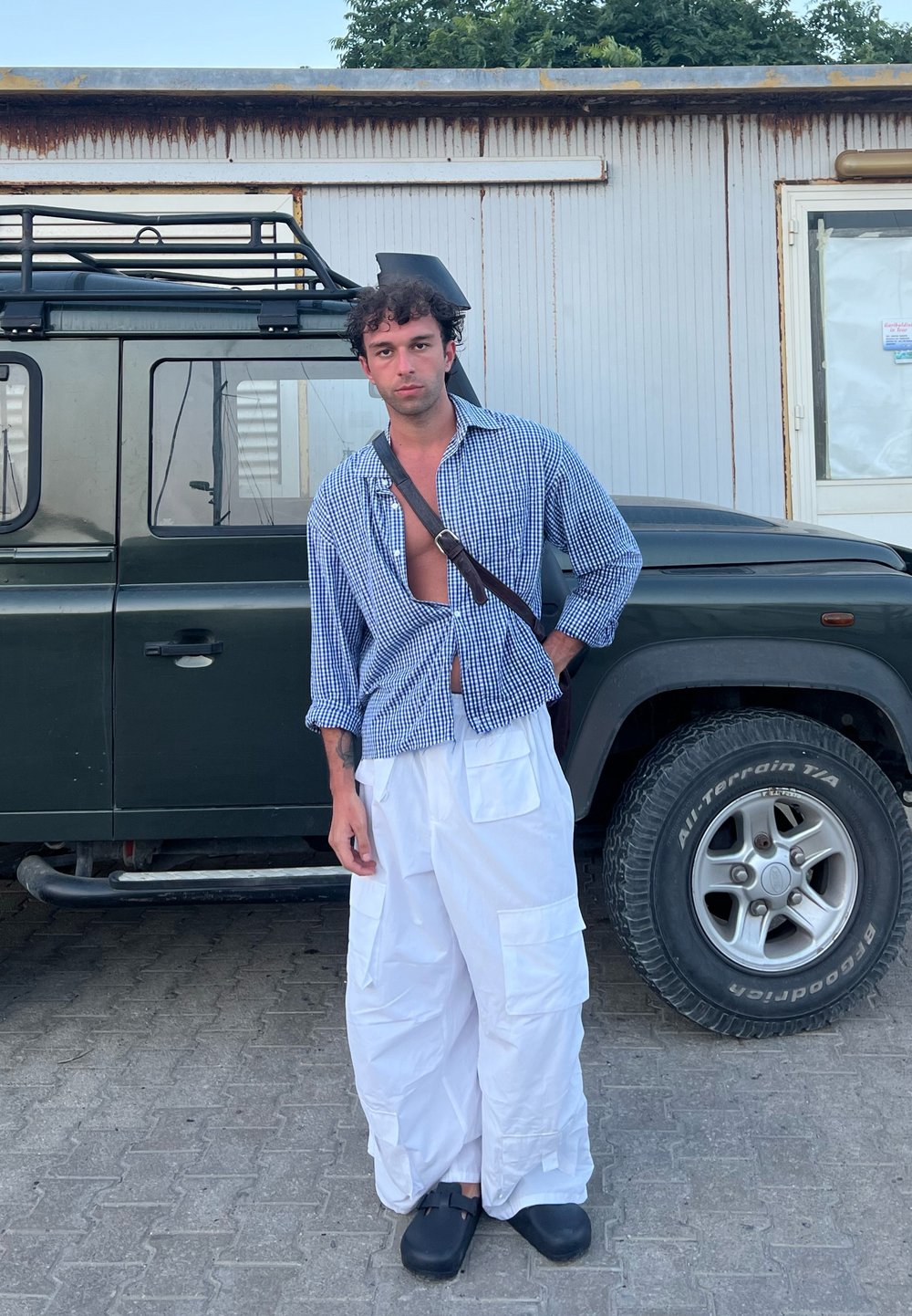 Avslappnad outfit bestående av en blå och vit rutig skjorta med öppen framsida, vita cargobyxor med fickor och svarta träskor, stående nära ett fordon.