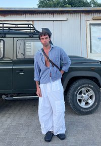 Avslappnad outfit bestående av en blå och vit rutig skjorta med öppen framsida, vita cargobyxor med fickor och svarta träskor, stående nära ett fordon.