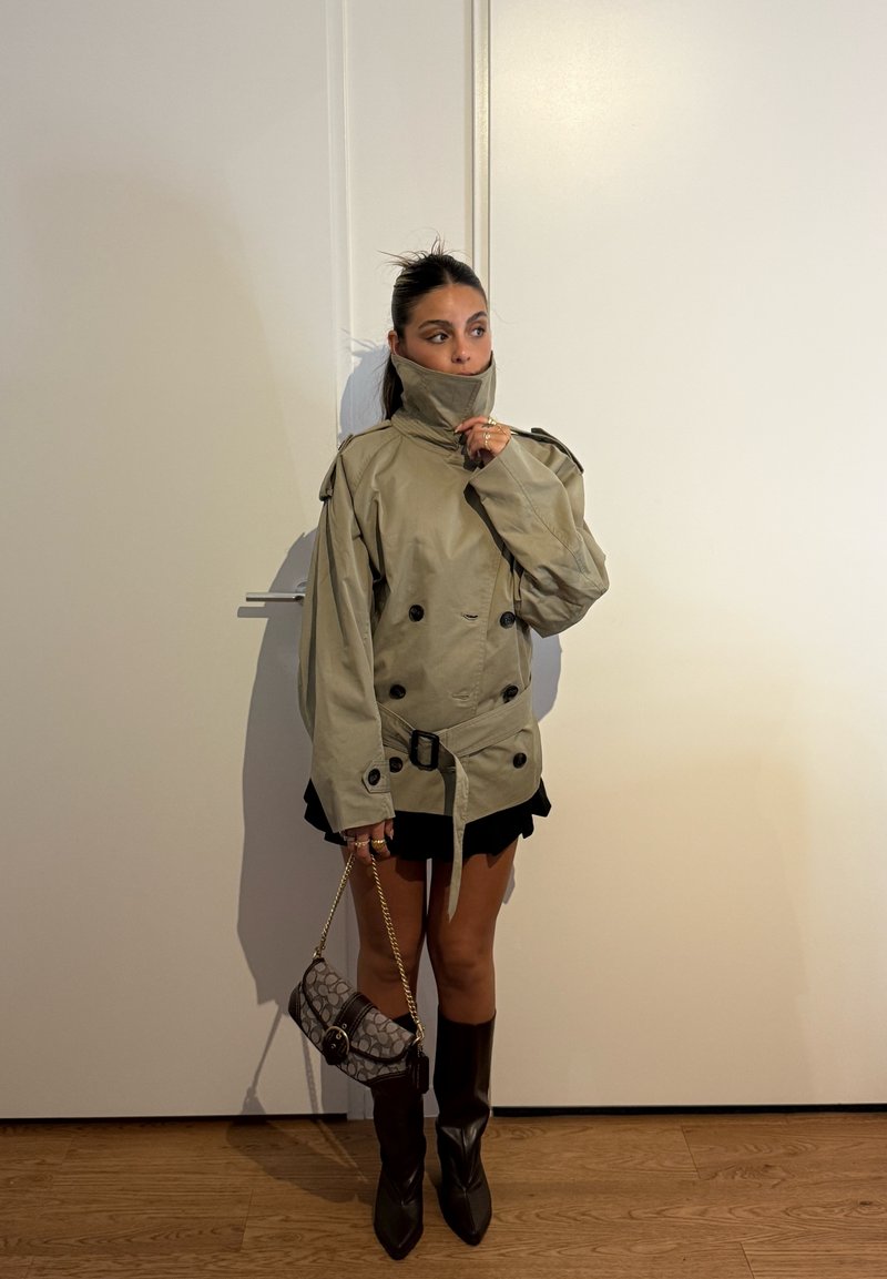 Femme debout à l'intérieur, portant un trench-coat beige surdimensionné avec un col haut, une jupe noire, des bottes noires jusqu'aux genoux, tenant un sac à main à motifs avec une chaîne.