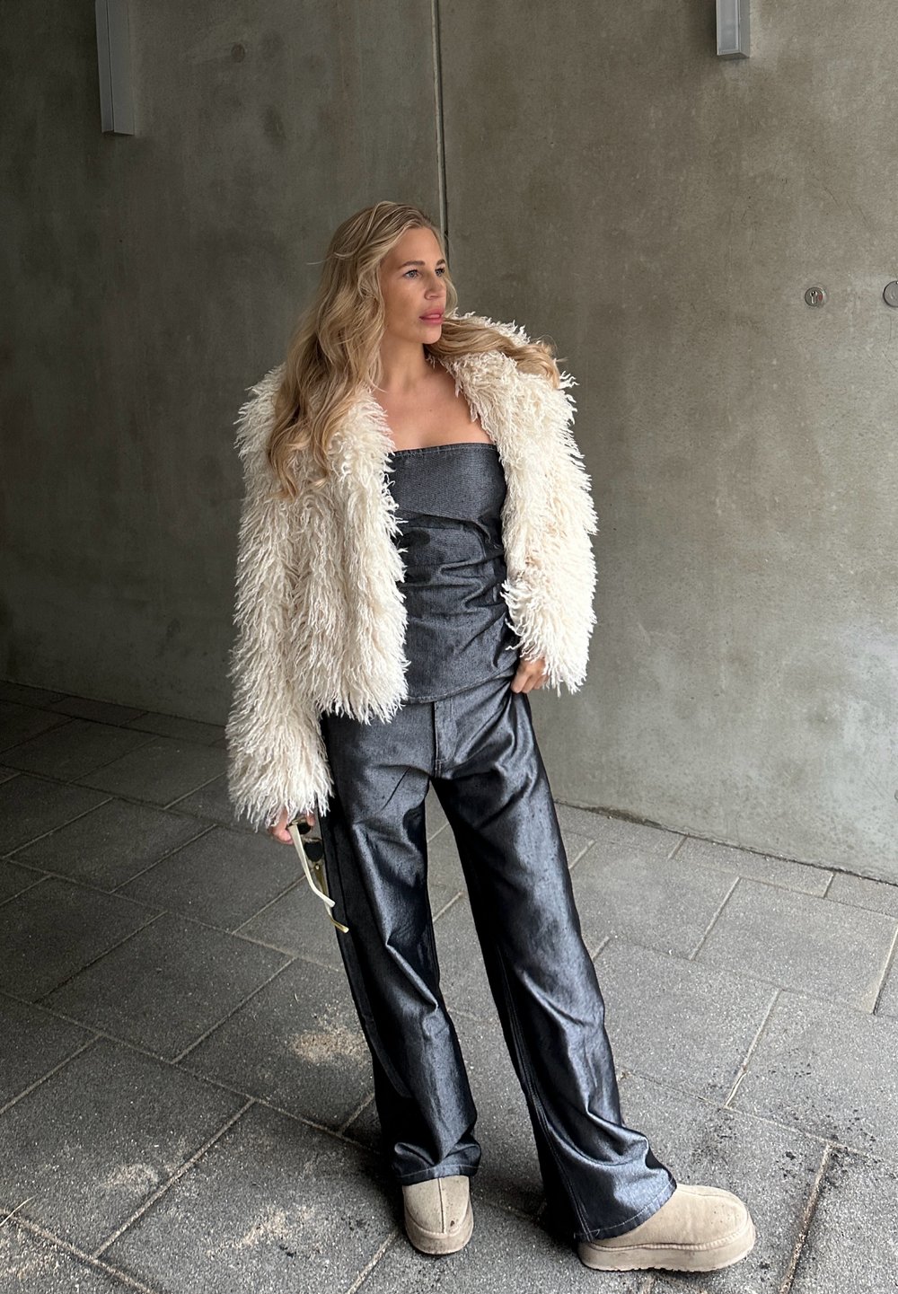 Veste en fausse fourrure crème, haut gris sans bretelles, pantalon large noir brillant, et bottines beiges. Lunettes de soleil tenues dans une main.
