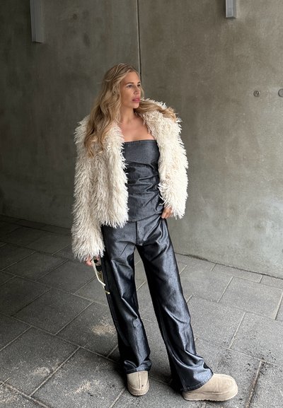 Chaqueta de piel sintética color crema, top gris sin tirantes, pantalones negros anchos y brillantes, y botines beige. Gafas de sol sostenidas en una mano.