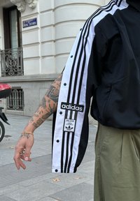 Black and white track jacket featuring a three-stripe design and snap fasteners. It includes an Adidas logo patch and is made from textured fabric.