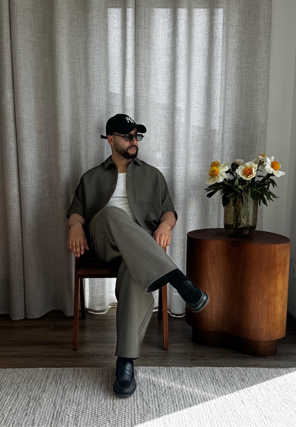 Uomo con cappello scuro, occhiali da sole, abbigliamento grigio, seduto su una sedia di legno accanto a un tavolo laterale in legno con un vaso di fiori bianchi e gialli.