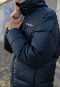 Casaco puffer preto com gola alta e fecho de correr. Apresenta um padrão acolchoado e o logo da Columbia. O tecido parece liso e isolado.