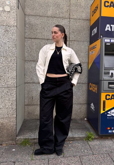 Chaqueta crema sobre top corto negro, pantalones anchos negros y zapatos negros. Sostiene un bolso negro texturizado con tachuelas. Fondo urbano con cajero automático.