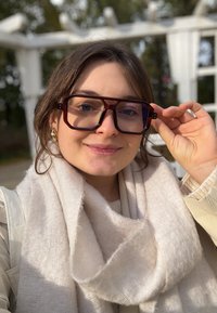 Ylisuuret suorakaiteen muotoiset silmälasit, joissa on tumman viininpunainen kehys ja kirkkaat linssit. Henkilöllä on luonnonmaisemassa vaalea huivi ja beige yläosa.