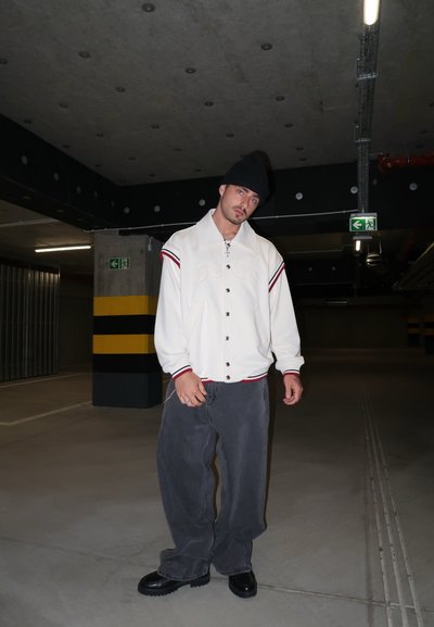 Chaqueta deportiva blanca con acentos y botones rojos, pantalones anchos grises y zapatos negros gruesos, situada en un garaje oscuro.