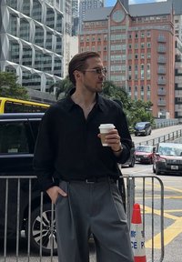 Camisa preta de manga comprida, calças largas cinzentas, segurando uma chávena de café. Usa óculos de sol e um colar prateado. Fundo urbano com veículos.