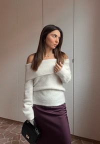 Maglione bianco a spalla scoperta in maglia con texture a coste, abbinato a una gonna di raso viola scuro e a una piccola borsa nera.