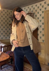 Beige corduroy jas over een bruine kabelgebreide trui, donkere spijkerbroek en een tan schoudertas; binnenomgeving met geruit behang.