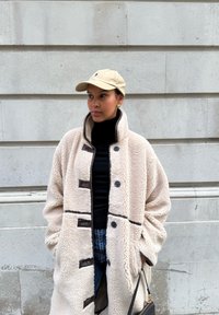 Manteau en fausse fourrure beige à col montant avec des accents foncés boutonnés, porté sur un col roulé noir et un jean bleu, assorti d'une casquette beige.
