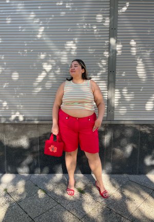 Top de tirantes a rayas, pantalones cortos rojos brillantes con bolsillos, sandalias rojas a juego y bolso con diseño floral, contra un fondo gris.