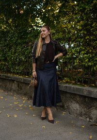 Hnedá kožená bunda, tmavá blúzka, deninová sukňa, hnedá kabelka, špicaté lodičky, stojí na chodníku s jesenným lístím, pozadie s zeleným porastom.