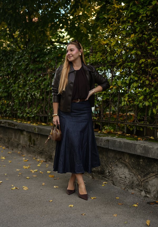 Jachetă din piele maro, bluză închisă, fustă din denim, poșetă maro, toc ascuțit, stând pe un alee cu frunze de toamnă, fundal de frunze verzi.