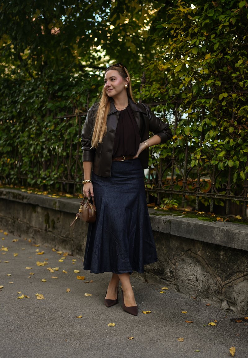 Brown leather jacket, dark blouse, denim skirt, brown handbag, pointed heels, standing on a pathway with autumn leaves, green foliage background.