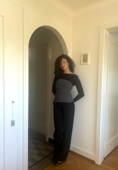 Una persona con un atuendo ajustado en gris y negro se encuentra en una puerta. La parte superior tiene mangas largas y un diseño estructurado; los pantalones son negros y de pierna ancha.