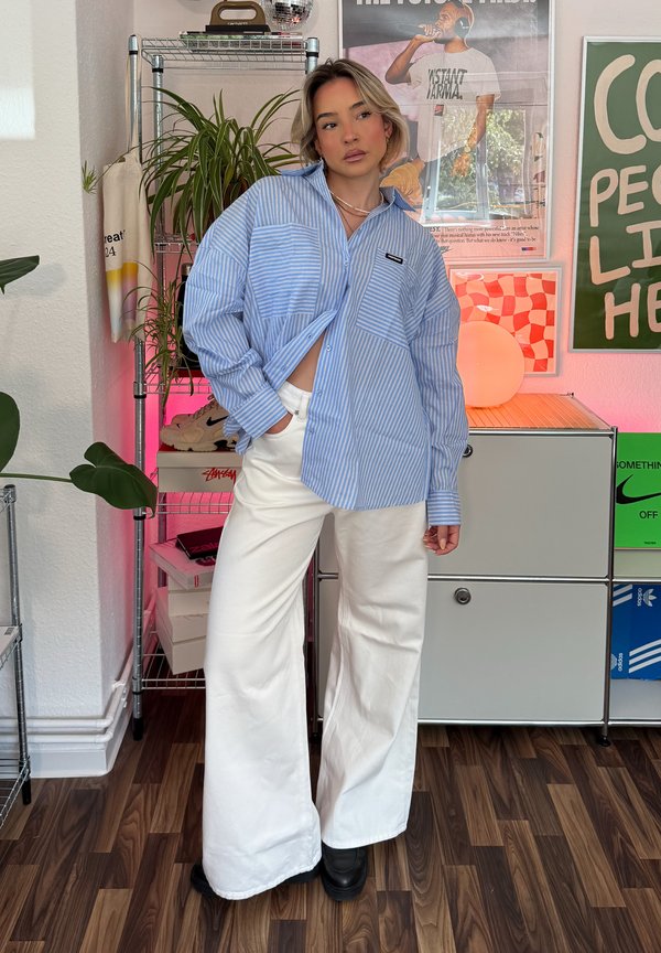 Blue and white striped button-up shirt featuring two chest pockets, teamed with white wide-leg trousers and black ankle boots. Set against a wooden floor backdrop.