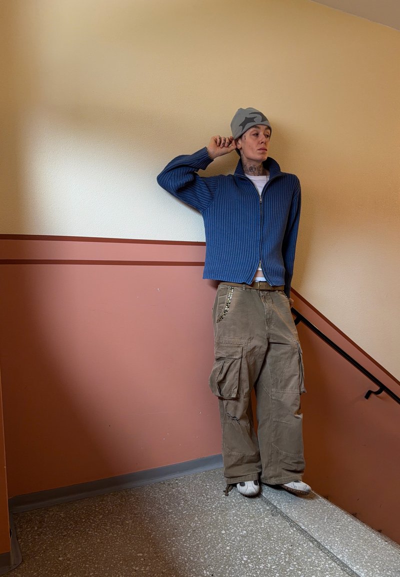 Jeune personne portant un bonnet gris, un pull zippé bleu et un pantalon cargo ample, debout dans un escalier intérieur devant un mur beige et marron.