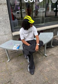 Homme portant une casquette jaune et noire, un t-shirt blanc et un pantalon noir, assis en tailleur sur un banc métallique courbe à l'extérieur d'un magasin.
