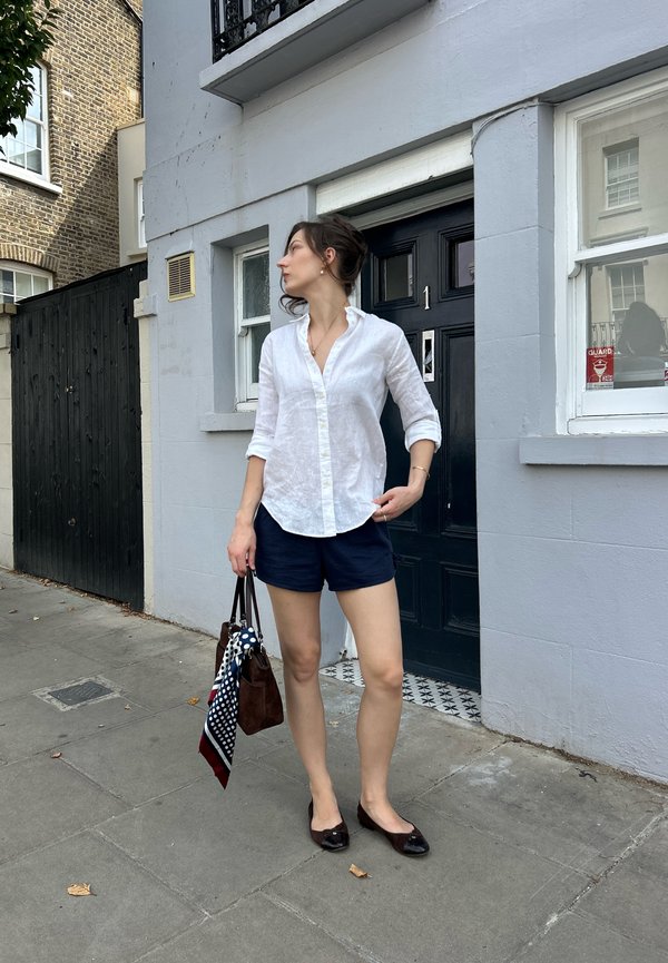 Mujer con camisa blanca y pantalones cortos azul marino está de pie en la acera sosteniendo un bolso marrón con una bufanda de colores cerca de una puerta negra con el número uno.