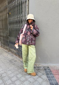 Puffy blomstrete jakke i mørke toner, lysegrønne quiltede bukser med lommer, beige bøttehatt, holder en kaffekopp, står nær en port.