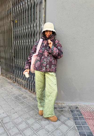 Chaqueta acolchada con estampado floral en tonos oscuros, pantalones acolchados verde claro con bolsillos, sombrero tipo pescador beige, sosteniendo una taza de café, de pie cerca de una puerta.