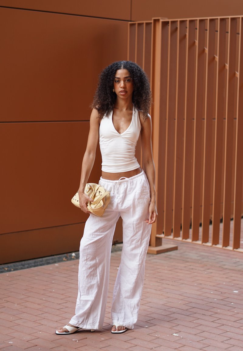 Femme aux cheveux bouclés portant un haut blanc sans manches, un pantalon blanc à jambes larges, des sandales blanches, tenant un sac à main beige, debout sur un chemin de briques.