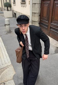 Person iført en sort dobbeltradet jakkesæt, hvid skjorte, ternet slips og mørke bukser, som holder en brun skuldertaske, med en sort kasket.