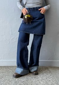 Jupe en denim superposée sur un pantalon large en denim, tous deux en indigo foncé. Grandes revers sur le pantalon avec un porte-clés texturé attaché à la taille.