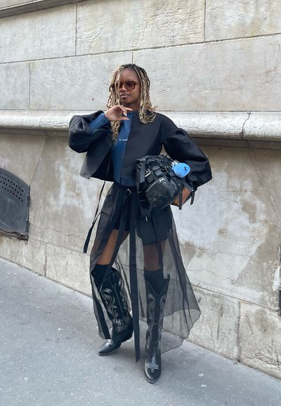 Chaqueta negra corta con mangas abullonadas, blusa azul, falda negra transparente con lazos y botas de cowboy negras hasta la rodilla con detalles en blanco. Bolsa negra.