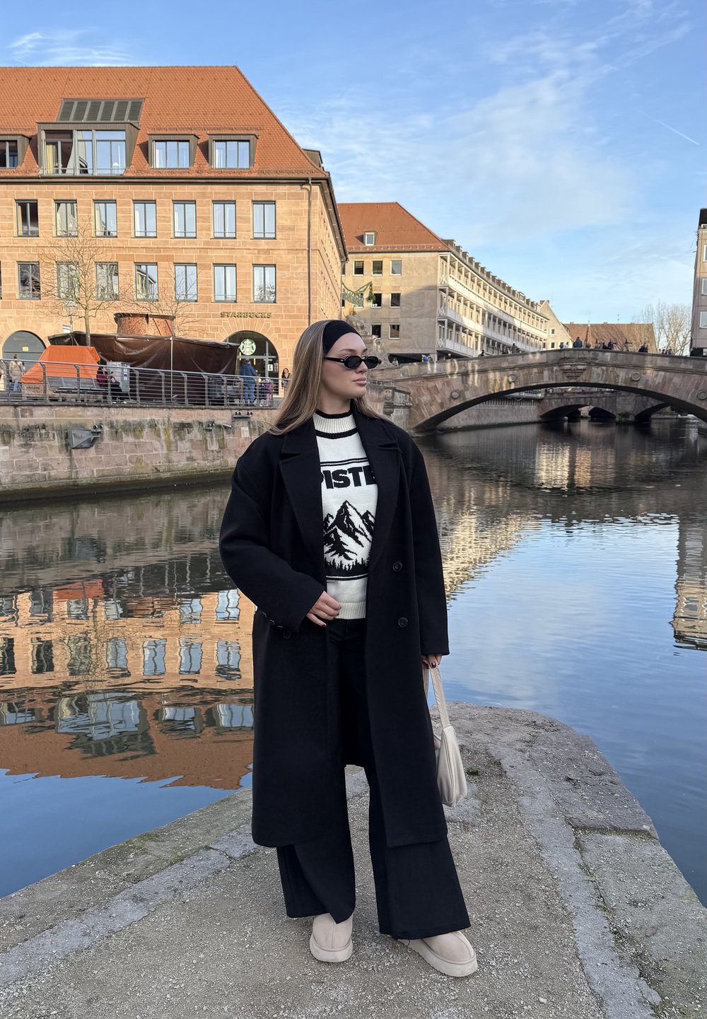 Femme portant un manteau noir et des lunettes de soleil, debout au bord d'une rivière avec un pont en pierre et des bâtiments aux toits en terre cuite sous un ciel bleu.